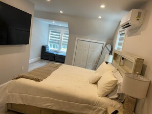 A bedroom with a bed, a bedside lamp, a TV mounted on the wall, a wardrobe, and a window with blinds.