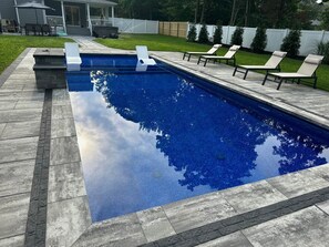 A rectangular swimming pool with a clear blue water surface, surrounded by a stone deck and equipped with white lounge chairs.