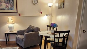 Un salon chaleureux avec un canapé gris, une petite table ronde ornée d’un vase de fleurs, un lampadaire et une horloge au mur.
