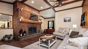 A living room with a brick fireplace, wooden beams, a ceiling fan, and a sofa. -  Private Adirondack Bear Retreat  (Athol)