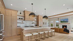 A modern kitchen with wooden cabinets, a central island with bar stools, pendant lighting, and a stone fireplace in the living area.
