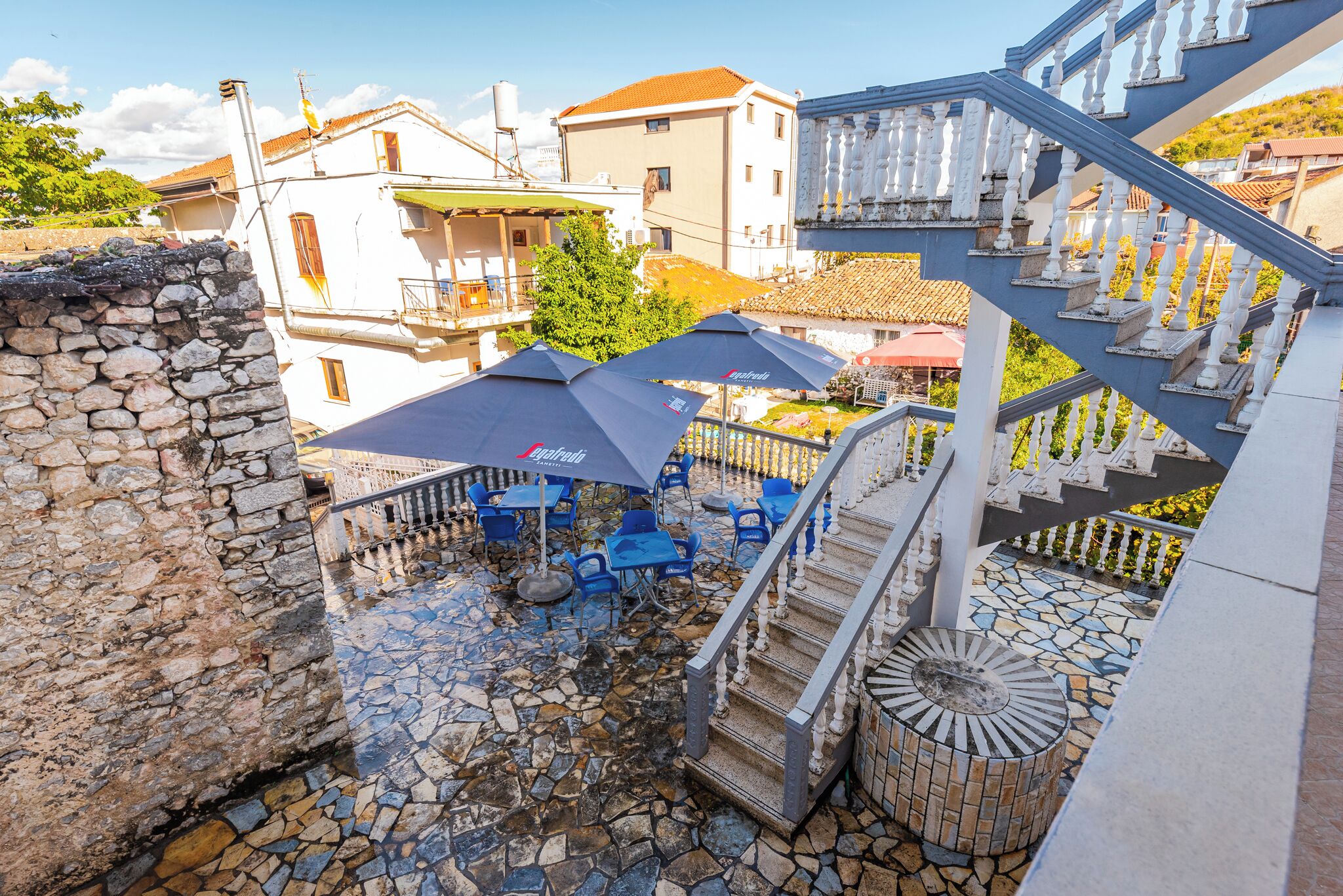 Un espace extérieur dallé de pierres, avec des chaises bleues et des parasols, un escalier et une vue sur les bâtiments environnants.