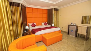 A hotel room with a large bed, an orange armchair, a desk with a mirror, and a minibar. - Apartment550 (Abuja)
