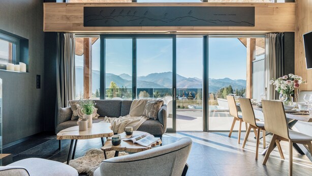 A modern interior with a large glass door leading to a mountain view. Inside, there is a sofa, a coffee table, and a dining area with chairs. - Wierchy View - Loft Affair Collection (Koscielisko)
