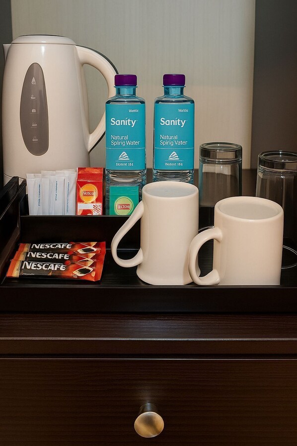 Un mini-bar d’hôtel contenant une bouilloire, deux bouteilles d’eau Sanity, deux sachets de café Nescafe et deux tasses blanches.