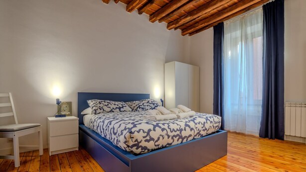 A bedroom with a bed, bedside table, chair, and a window with curtains. - Cozy Flat Orso Navona square (Roma)