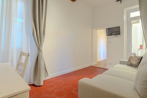 A room with a white table, a white chair, a sofa, a TV mounted on the wall, and a window with curtains. - Cozy Flat Orso Navona square (Roma)