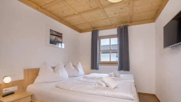 A bedroom with a wooden ceiling, a bed with white bedding, a bedside table with a lamp, and a TV mounted on the wall. - Austrian Mountain Chalet Retreat (Wald im Pinzgau)
