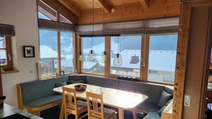 A cozy room with a wooden ceiling, a dining table with chairs, a bench, and large windows offering a view of snow-covered roofs. - Austrian Mountain Chalet Retreat (Wald im Pinzgau)