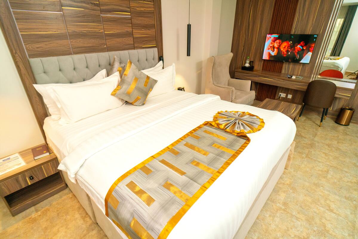 A neatly made bed with a patterned bedspread, a headboard with a wood-like design, a television on the wall, and a desk with a chair.