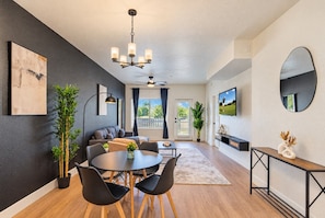 A modern living room with a dining area, a large window, a ceiling fan, and a flat-screen TV.