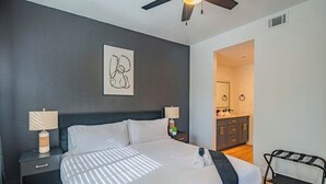 A neatly made bed with a headboard, two bedside tables with lamps, a wall-mounted artwork, and a glimpse of a bathroom and kitchen area.