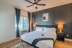 Une chambre à coucher comprenant un lit, deux lampes de chevet, un ventilateur de plafond, une fenêtre avec des rideaux et une œuvre d’art accrochée au mur.