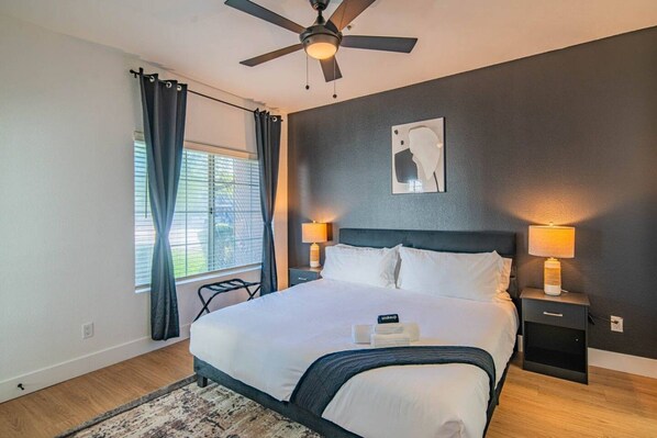 A bedroom with a bed, two bedside lamps, a ceiling fan, a window with curtains, and a wall-mounted artwork.