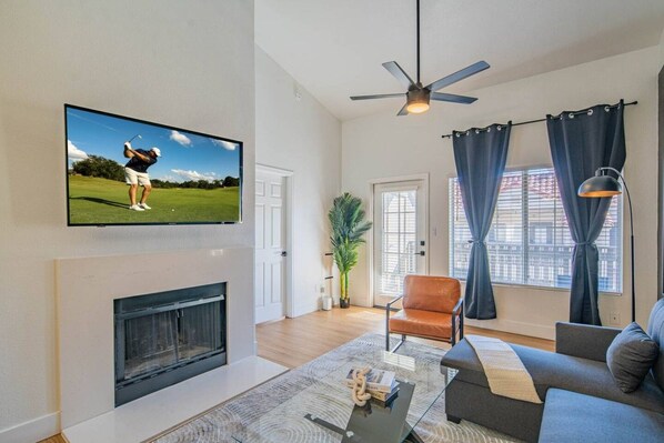 A living room with a flat-screen TV mounted on the wall, a fireplace, a ceiling fan, a sofa, a coffee table, and a window with curtains.