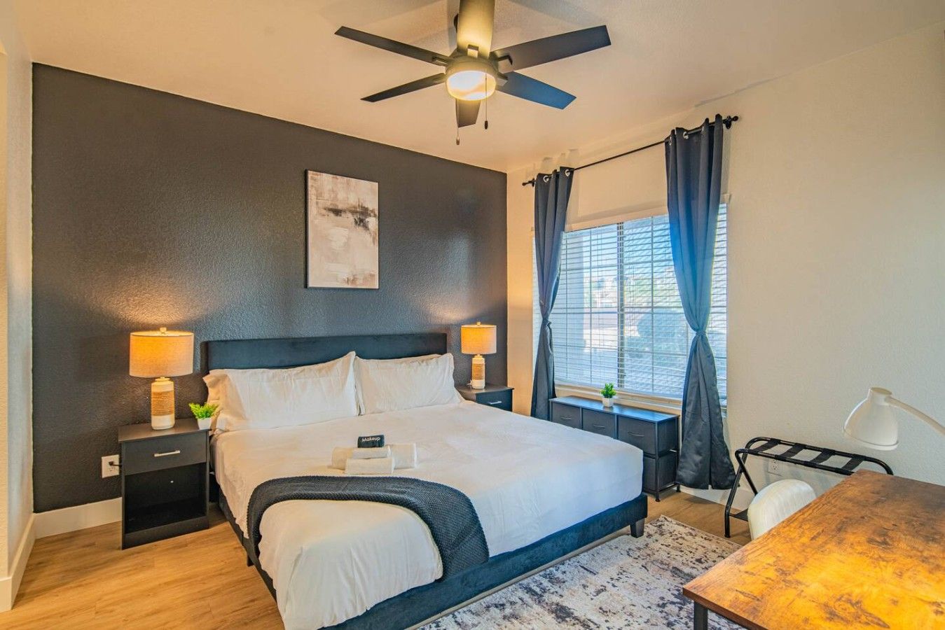 A modern bedroom with a bed, bedside lamps, a ceiling fan, a window with curtains, and a desk with a chair.
