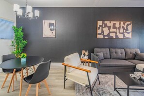 Ein modernes Wohnzimmer mit dunkler Wand, einem grauen Sofa, einem runden Tisch mit Stühlen und einer kleinen Pflanze.