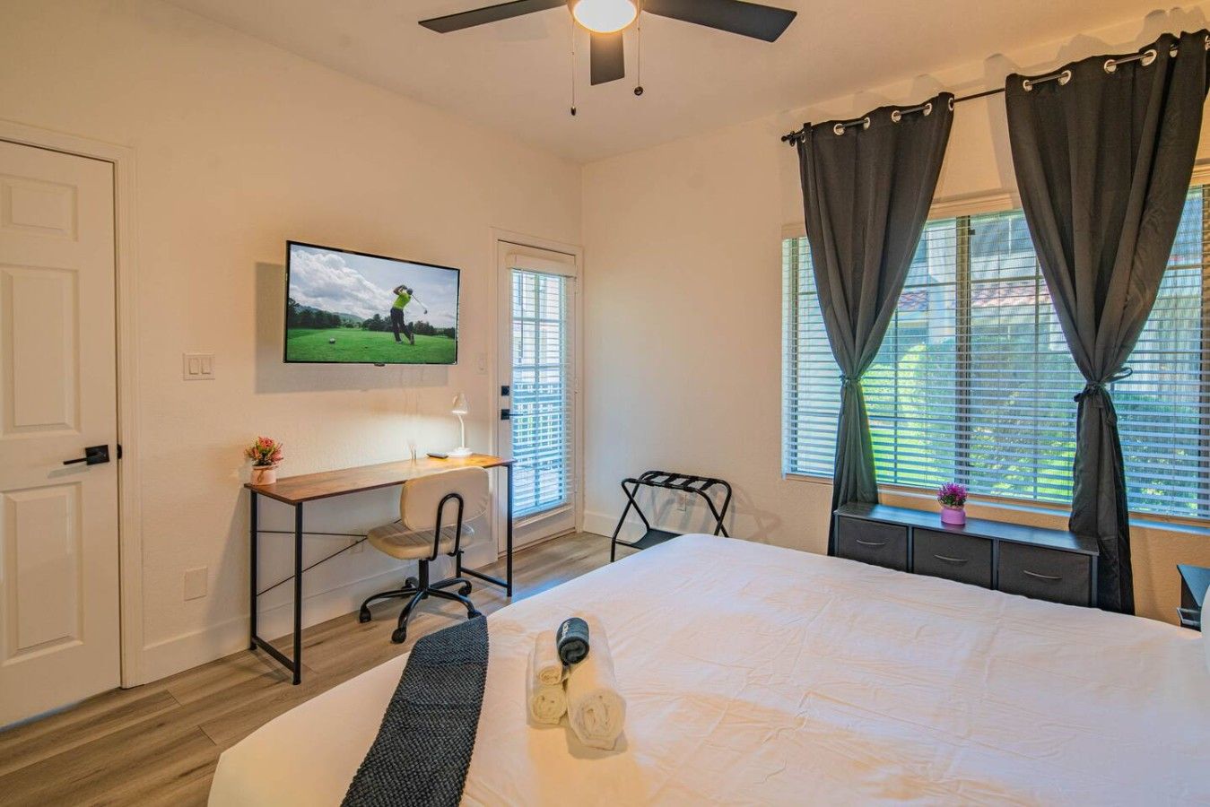 Une chambre à coucher avec un lit, un bureau, une chaise, un téléviseur fixé au mur et une fenêtre avec des rideaux.