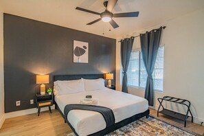 Ein Hotelzimmer mit einem Bett, zwei Nachttischlampen, einem Deckenventilator, einem Fenster mit Vorhängen und einem kleinen Tisch mit einer Pflanze.