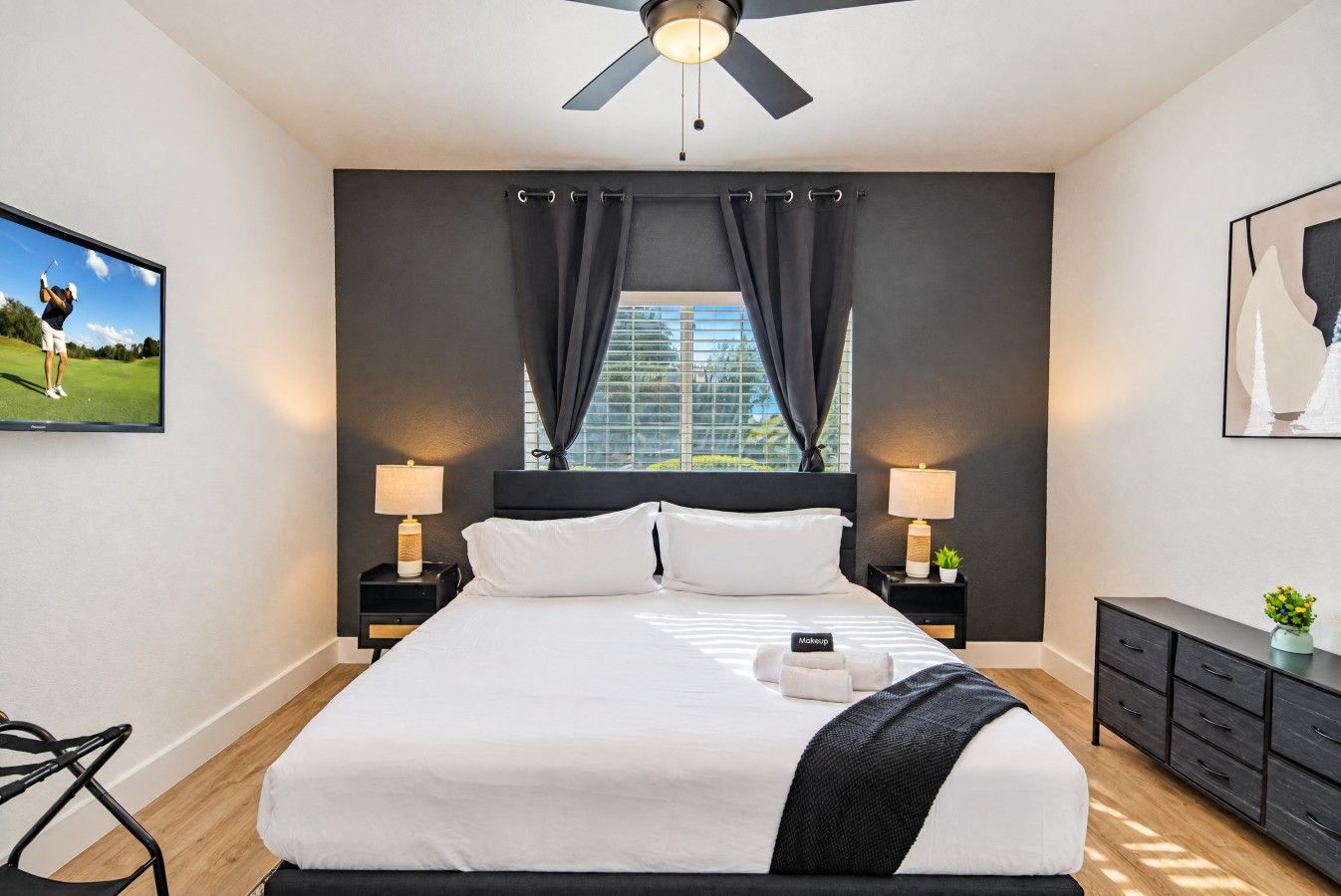 A modern hotel room with a large bed, two bedside tables with lamps, a ceiling fan, a window with curtains, a framed picture on the wall, and a small plant on a dresser.