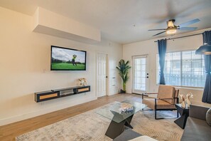 A modern living room with a flat-screen TV mounted on the wall, a glass coffee table, a comfortable sofa, and a ceiling fan.