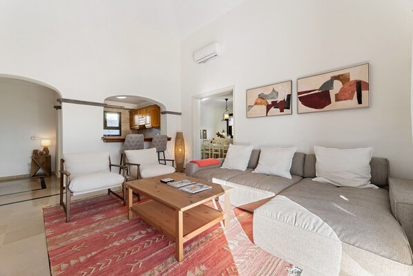 Una sala de estar moderna con un sofá, sillones, una mesa de centro de madera y una alfombra. Se ve una cocina al fondo.