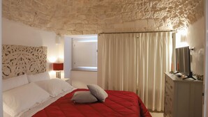 Une chambre avec un plafond en pierre, un lit avec une literie blanche et rouge, une commode avec un miroir et une fenêtre avec des rideaux.