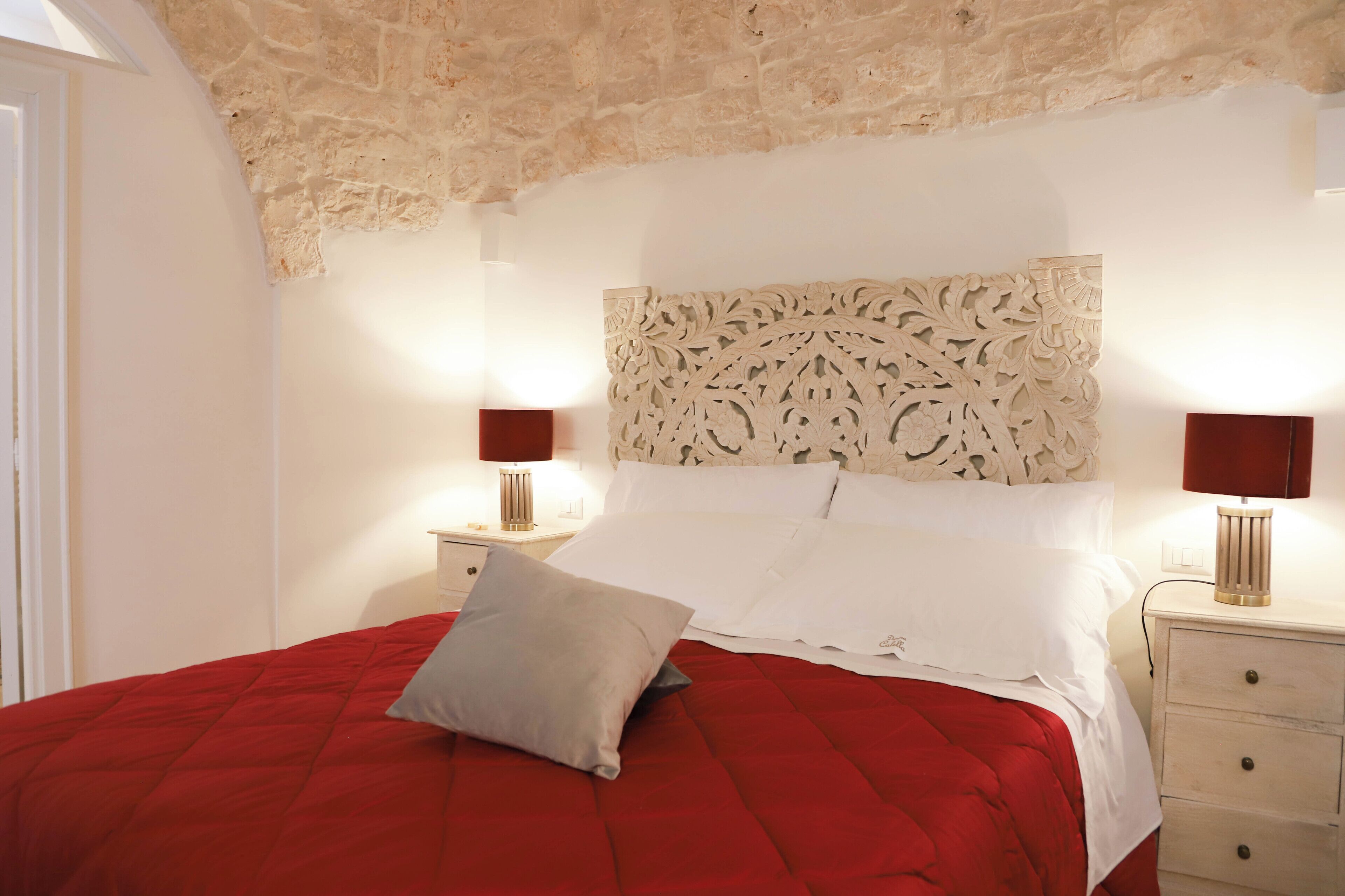 A bed with a red quilt, white pillows, and a decorative headboard. There are two bedside tables with lamps.