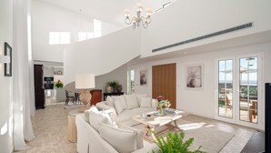 A spacious living area with a large sofa, a coffee table, and a chandelier. There is a kitchen in the background and a view of the outdoors through large windows.