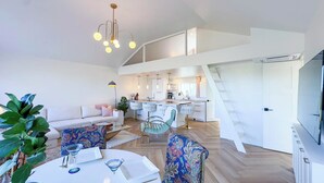 A modern living room with a sofa, a dining table, and bar stools. There is a kitchen area with a bar and a staircase leading to an upper level. - Modern Classic, Lake Views (Kincardine)