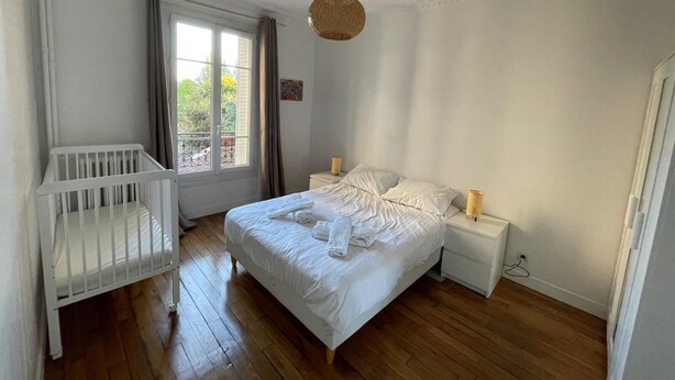 A bedroom with a wooden floor, a white crib, a bed with white linens, a bedside table with a lamp, and a window with curtains. - Apartment in Gentilly, 2 steps from Paris (Gentilly)