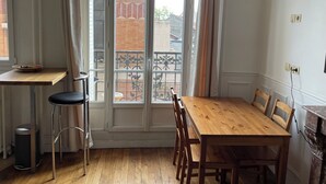 A room with a wooden dining table, chairs, a small stool, a trash bin, and a window with a view of a balcony and buildings. - Apartment in Gentilly, 2 steps from Paris (Gentilly)