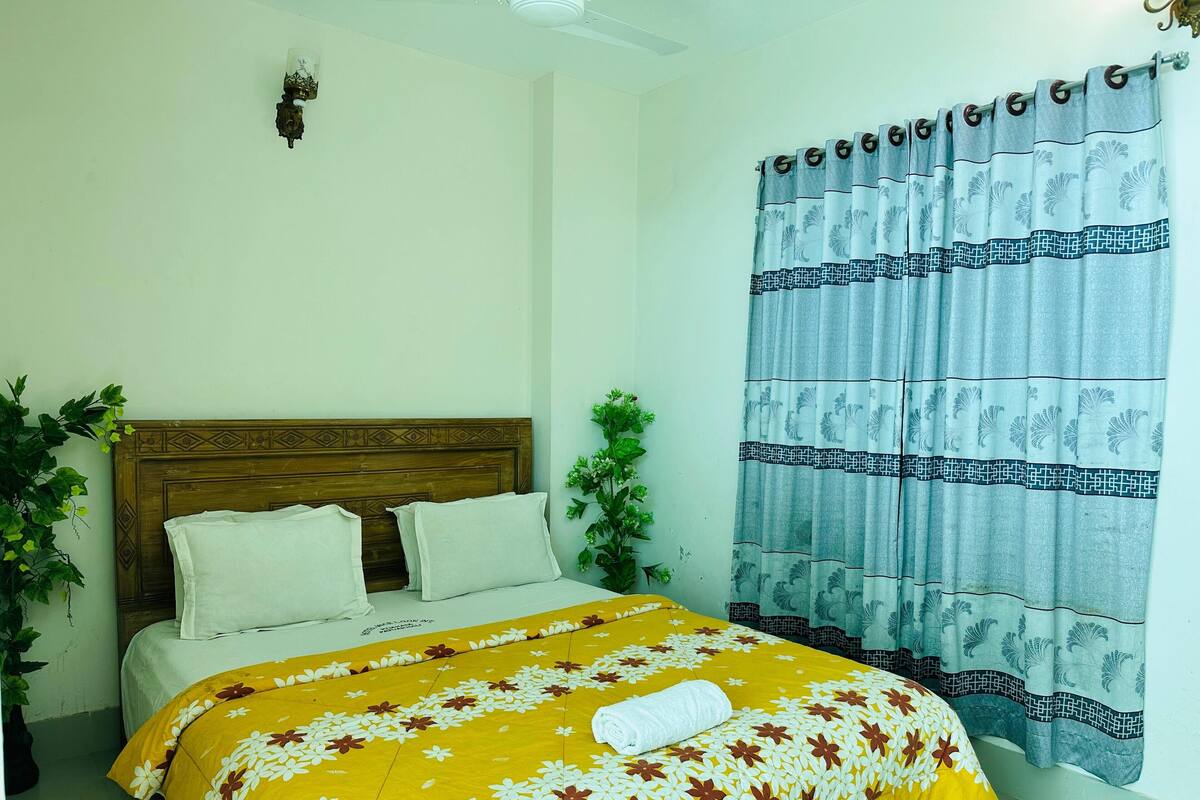 A bedroom with a wooden headboard, a bed with a yellow floral bedspread, white pillows, and a rolled white towel. There is a potted plant on the left and a curtain with a leaf pattern on the right.