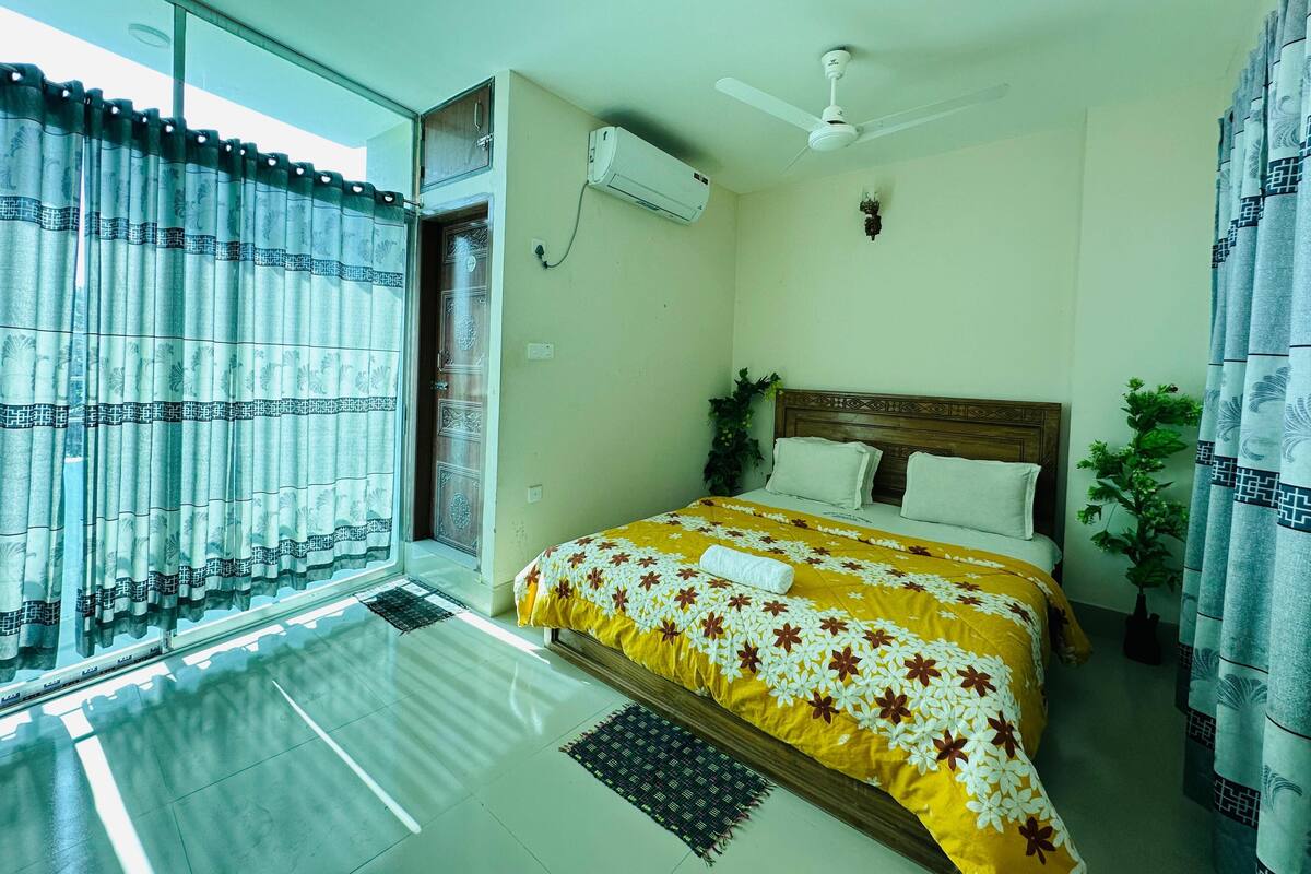 A bedroom with a bed, a ceiling fan, an air conditioning unit, and a window with curtains.