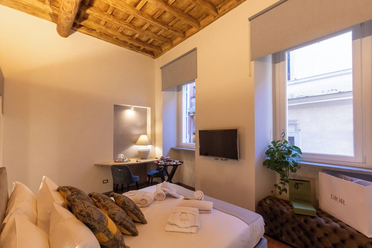 A hotel room with a wooden ceiling, a bed with neatly folded towels, a desk with a lamp, a TV, and a window with a plant and shopping bags.