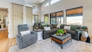 A spacious living room with a gray sofa set, a small side table, and a coffee table. There are large windows providing natural light and a view of the outdoors. - Dreamy Hilltop View: Roseburg Wine Country Retreat (Roseburg)