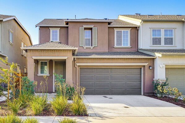 A two-story residential building with a garage, a small covered entryway, and a landscaped front yard with various plants.