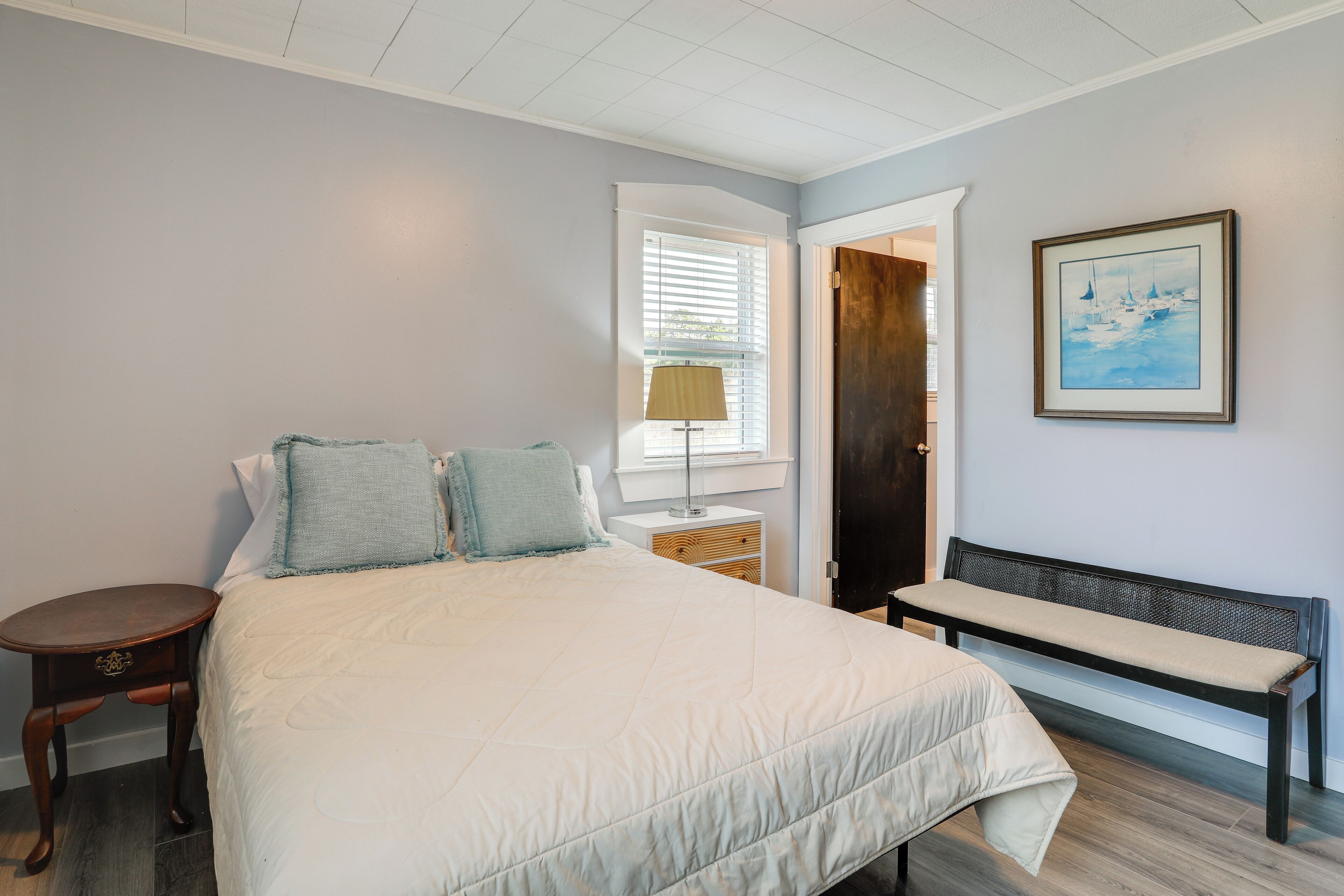 A bedroom with a large bed, a bench, a nightstand, a lamp, a door, and a framed picture on the wall.