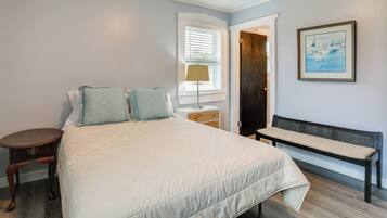 A bedroom with a large bed, a bench, a nightstand, a lamp, a door, and a framed picture on the wall.