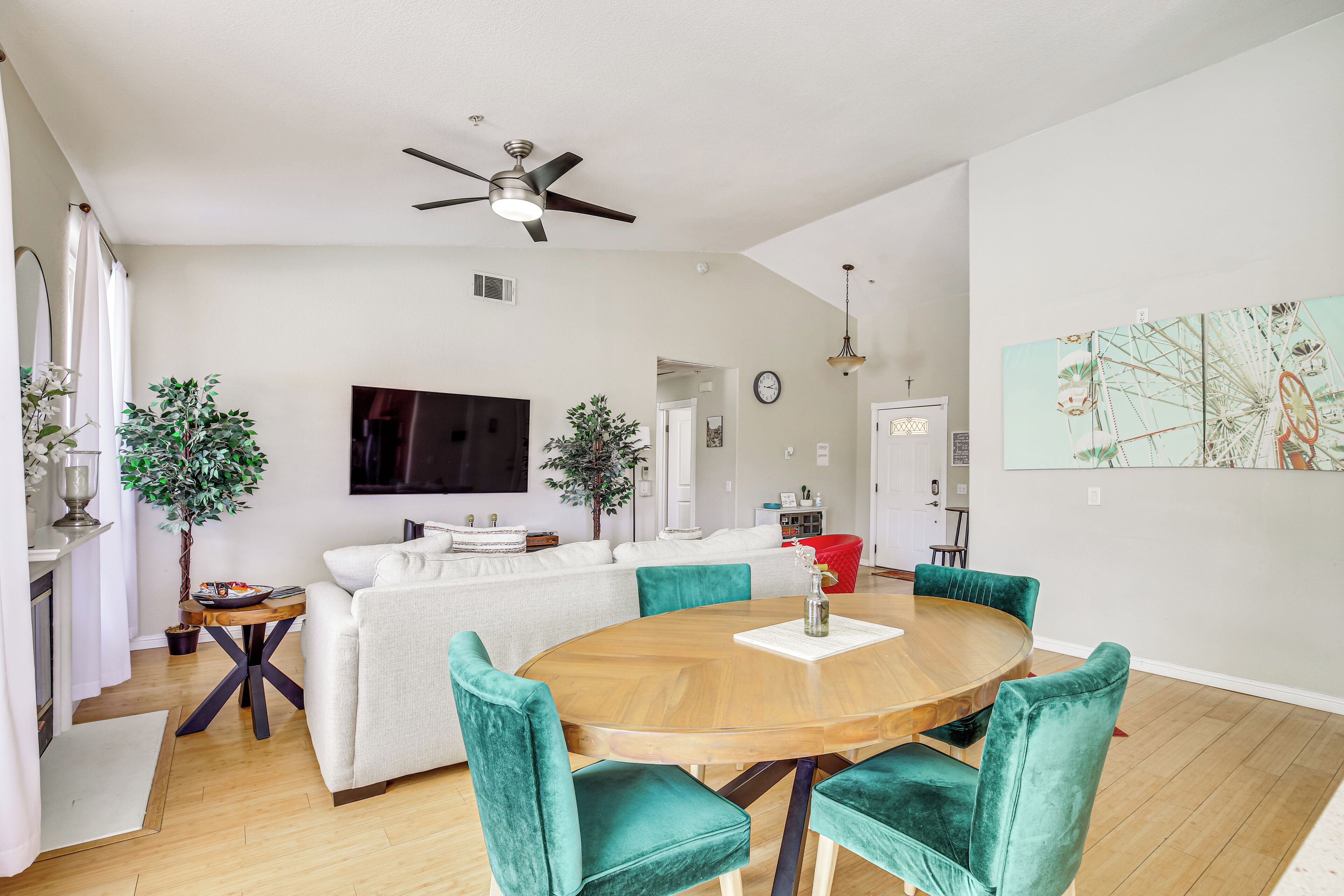 A modern living room with a round wooden dining table, teal upholstered chairs, a white sofa, a flat-screen TV, and a ceiling fan.