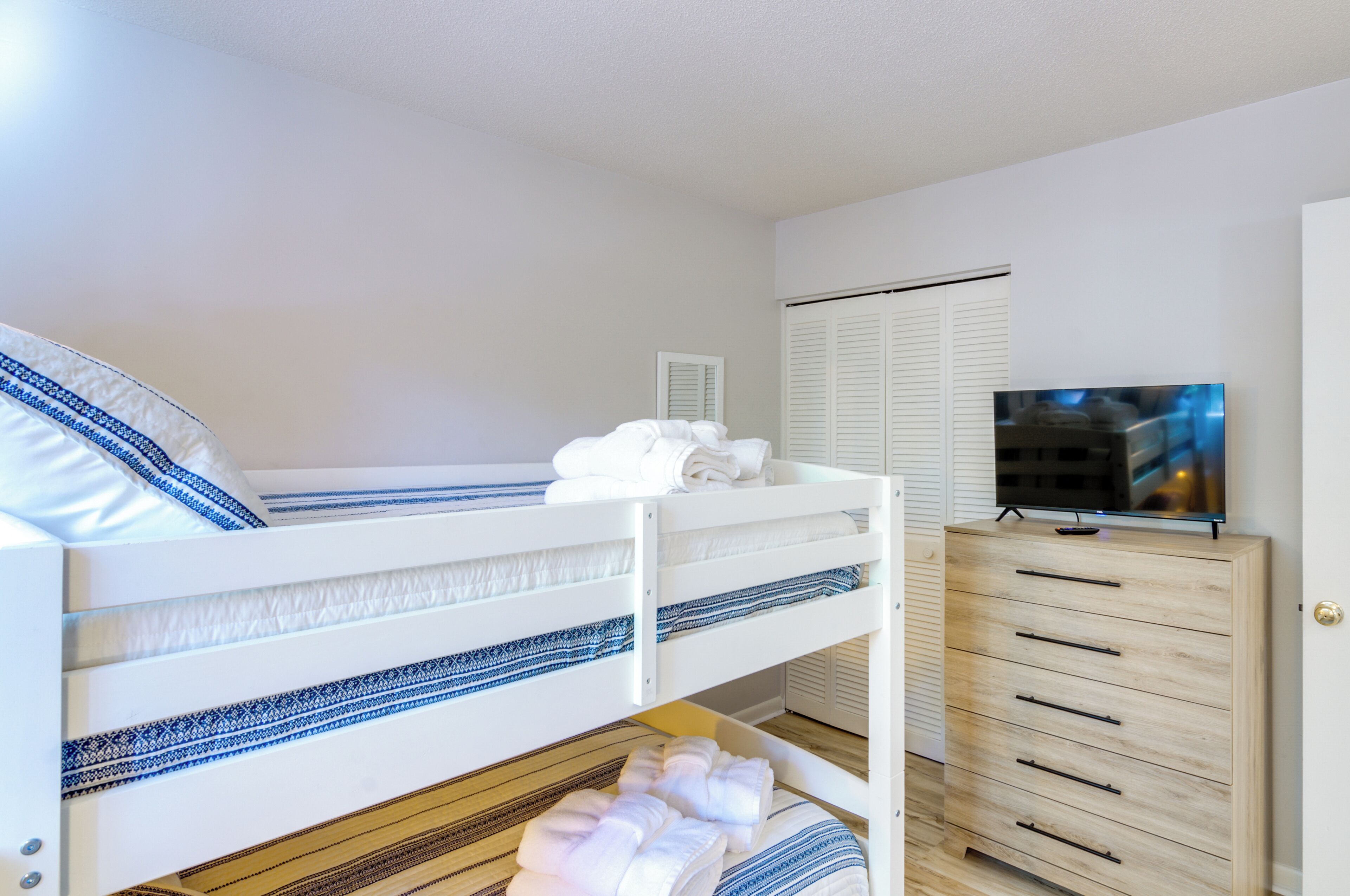 A bunk bed with white bedding and blue patterns, a wooden dresser with drawers, and a television on a stand.