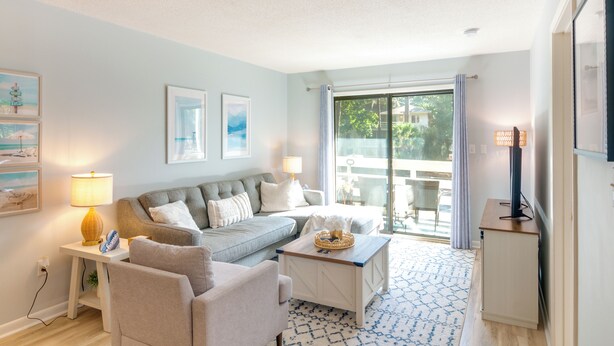 A modern living room with a sofa, armchairs, a coffee table, and a TV. There is a large window with a view of greenery outside. - Walk to Beach: Condo w/ Balcony in Hilton Head! (Hilton Head Island)