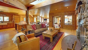 A spacious living area with wooden floors, a stone fireplace, and a large window overlooking a scenic landscape. - Sun Valley Log Home w/ Hot Tub & Mtn Views! (Sun Valley)
