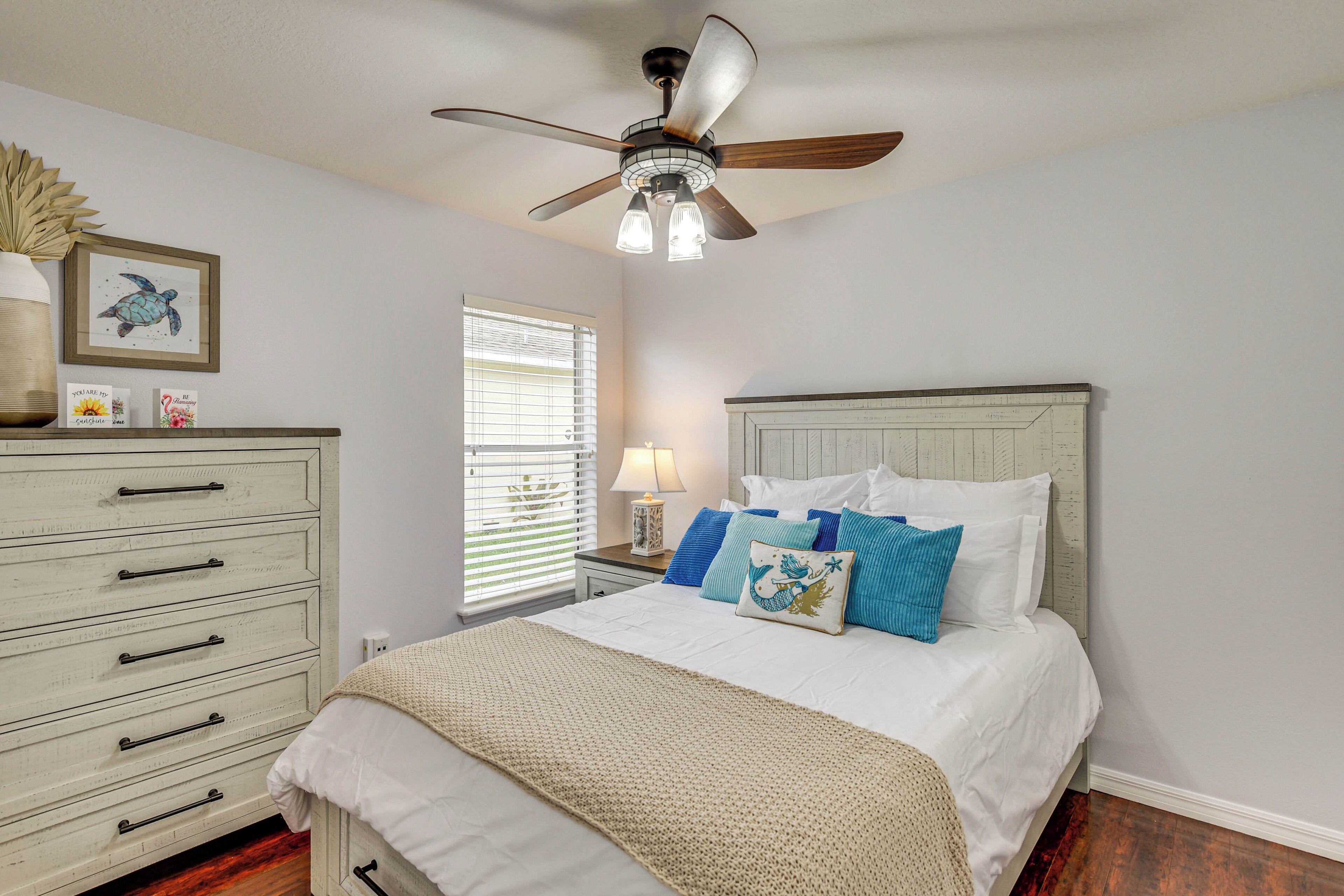 A bedroom with a bed, dresser, ceiling fan, and a window with blinds.