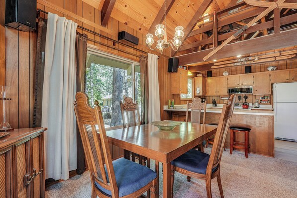 A wooden dining table with chairs, a chandelier, and a kitchen area with cabinets and a refrigerator.