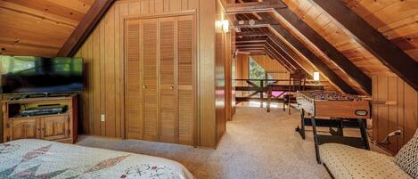 A cozy attic bedroom with a wooden ceiling, a bed with a floral quilt, a television, and a foosball table.