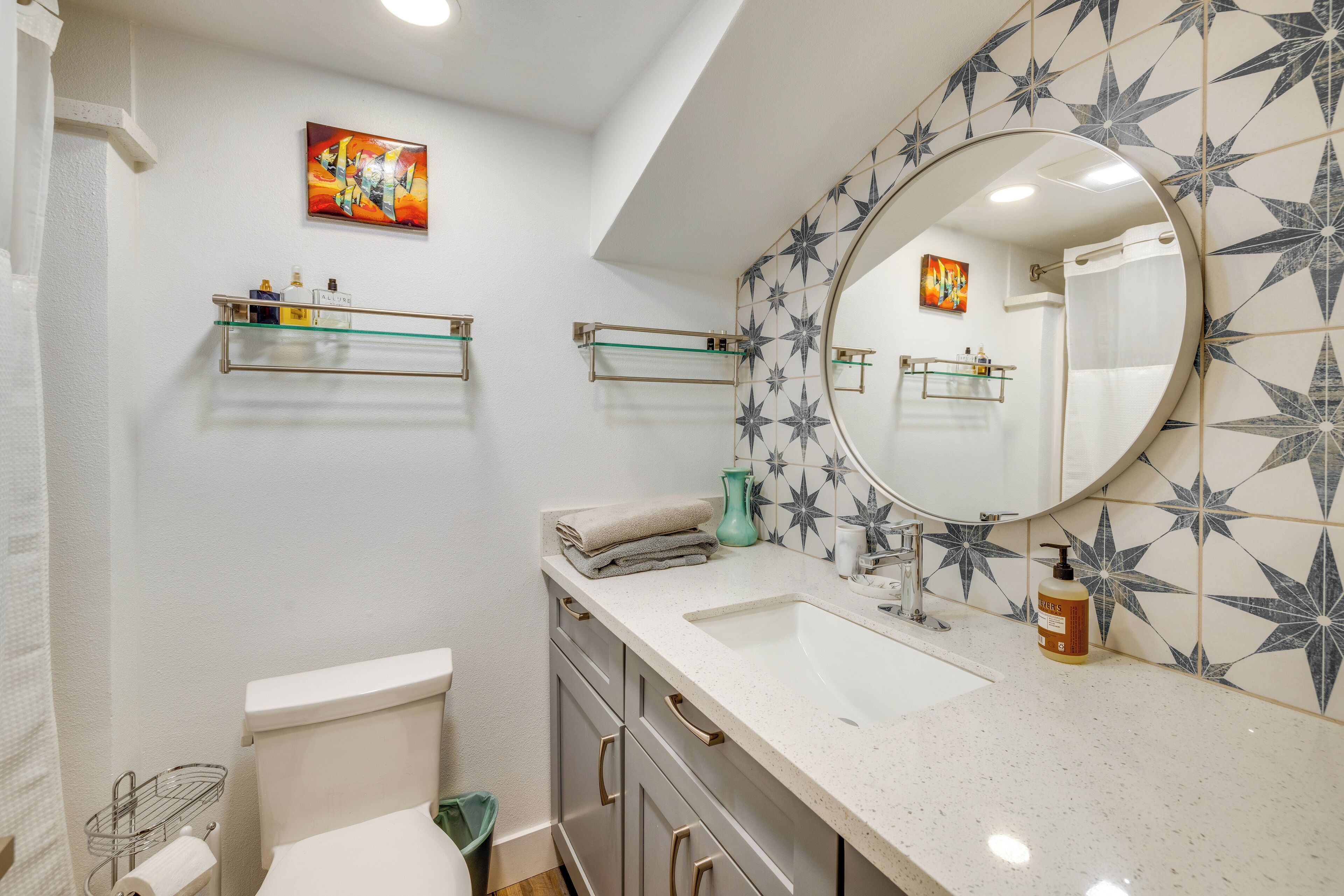 A bathroom with a white toilet, a sink with a mirror, and a shower curtain. There are two shelves with toiletries and a decorative tile pattern on the wall.
