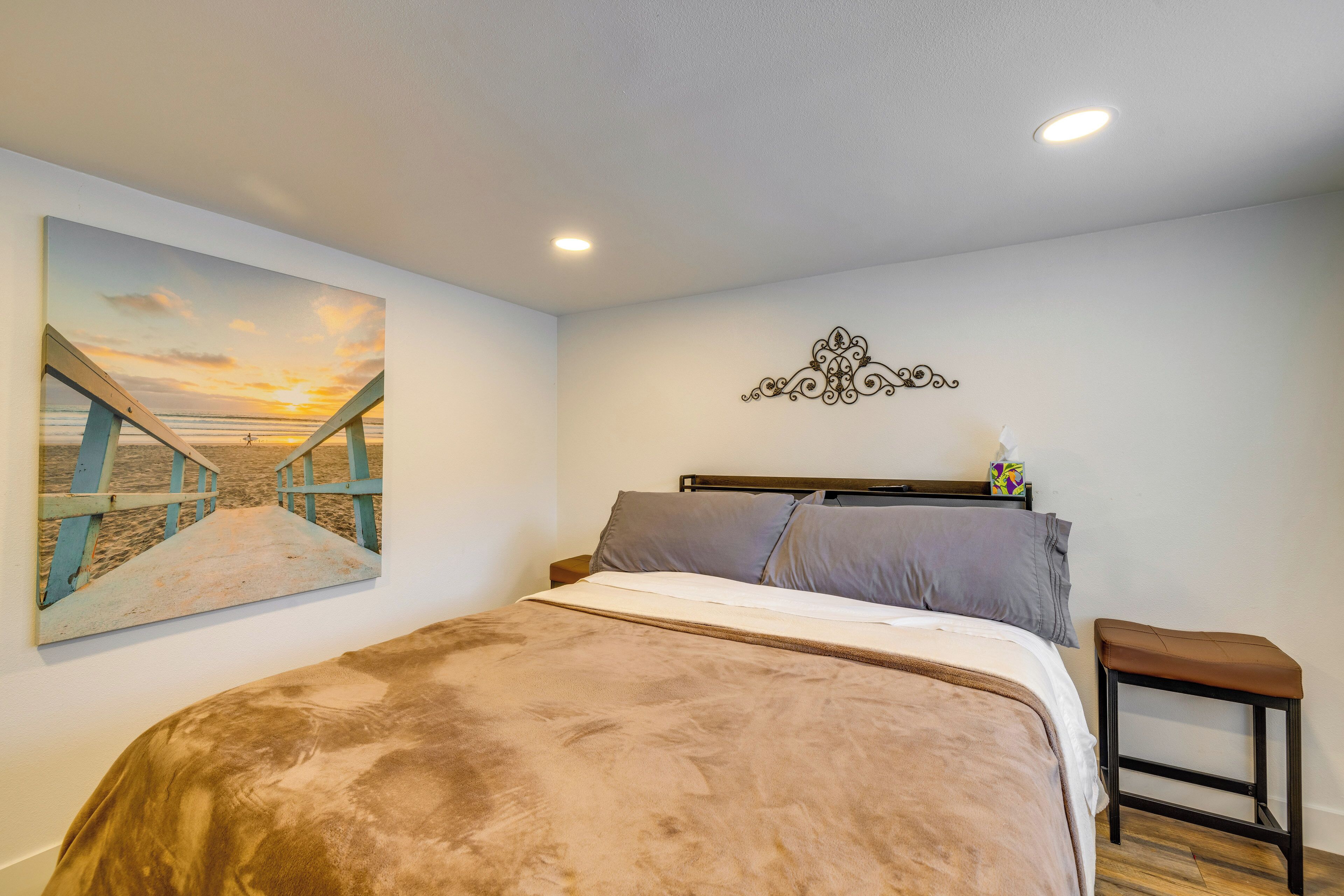 A bedroom with a large bed, a nightstand, a wall-mounted decorative piece, and a painting of a sunset over a bridge.