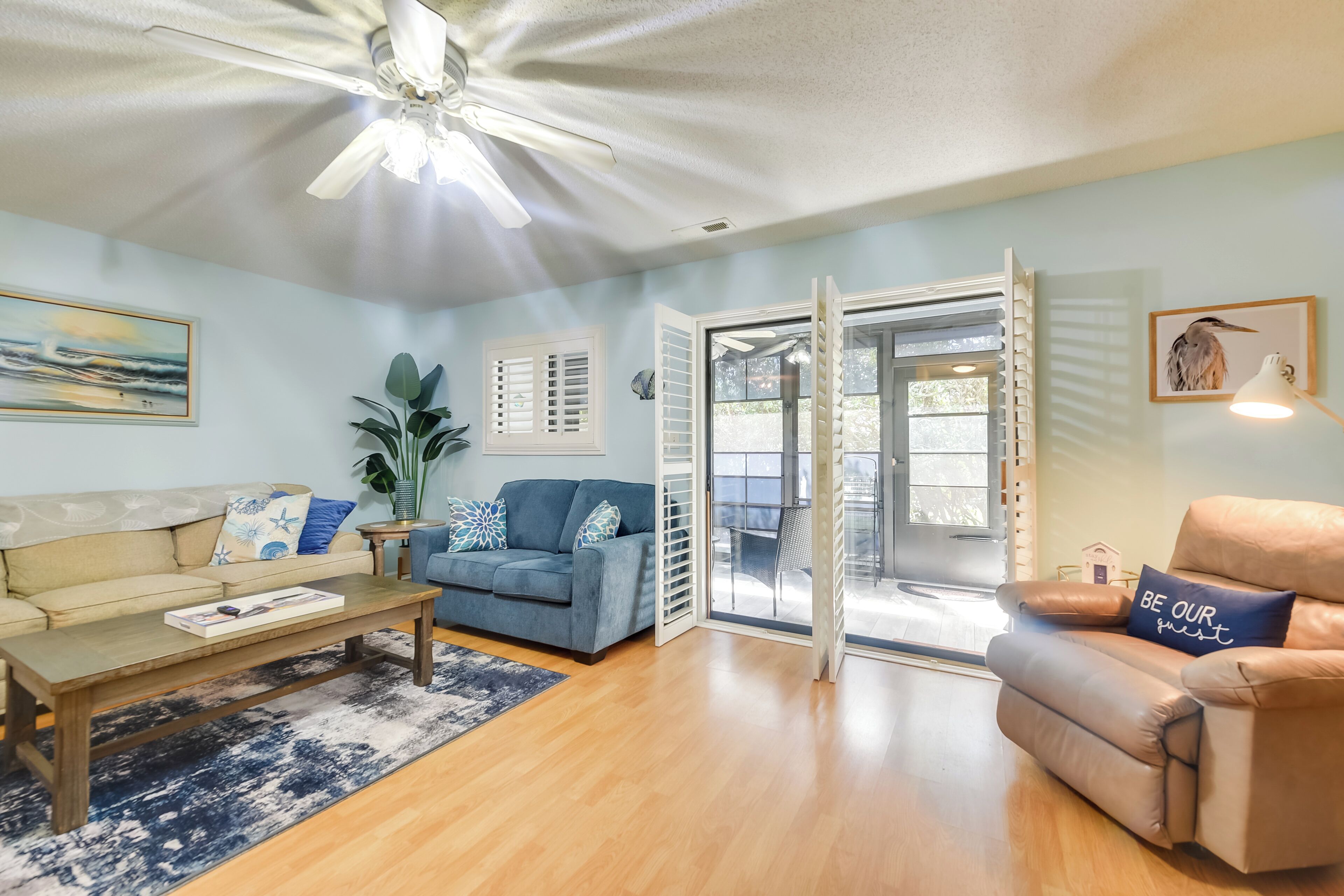 A living room with a sofa, a coffee table, a blue couch, a brown armchair, a ceiling fan, and a sliding glass door leading to an outdoor area.