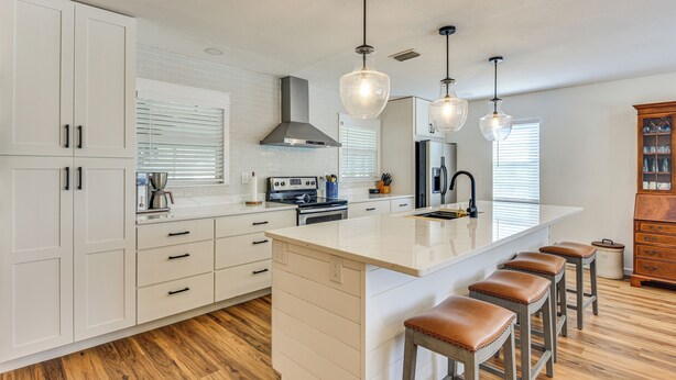 A modern kitchen with white cabinetry, a large island with bar stools, a stainless steel range hood, and pendant lighting.